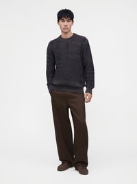 Homme debout portant un pull en maille texturée foncé, un pantalon marron ample et des baskets marron sur fond blanc uni.
