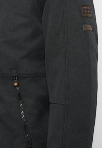 Tmavě šedá bunda vyrobená z texturované látky. Disponuje zipovou kapsou, ramenními nášivkami a kontrastním oranžovým táhlem na zip.