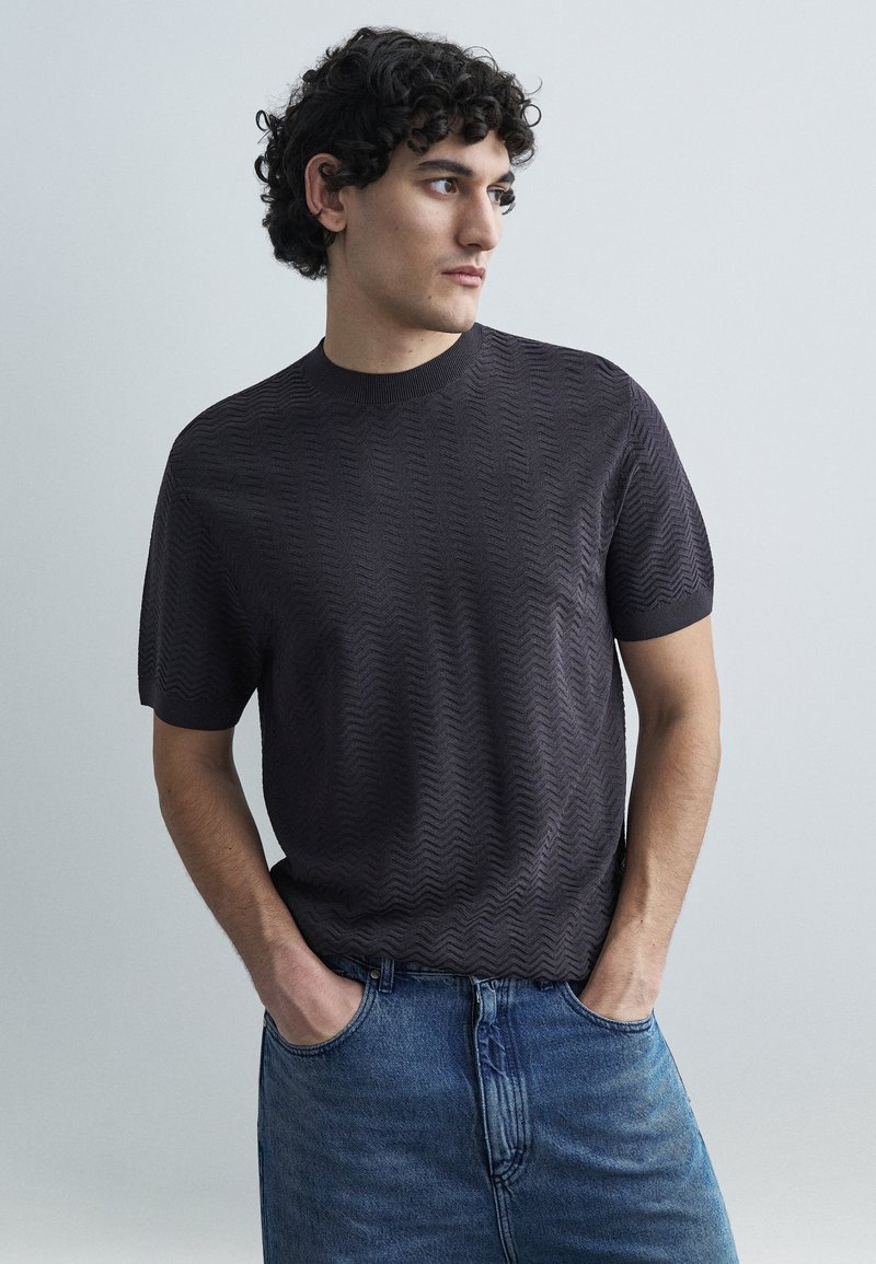 Tričko s krátkym rukávom v tmavosivej farbe s textúrou a okrúhlym výstrihom, doplnené modrými džínsami. Tričko má jemný zygzakovitý vzor.