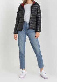 Kvinna som bär en svart puffjacka med röd foder, en ljusgrå tröja, blå högmidjade jeans och vita sneakers stående mot en enkel bakgrund.
