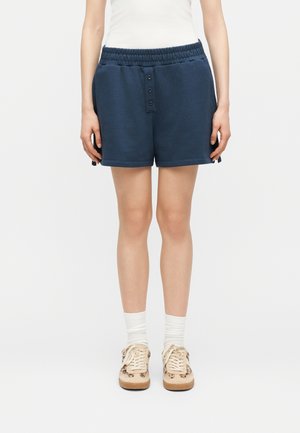 Person iført mørkeblå elastiske shorts med knapdetaljer, hvide sokker, beige sneakers og en hvid ærmeløs top, stående oprejst.
