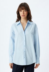 Camicia azzurra chiaro con bottoni, maniche lunghe, colletto classico e vestibilità rilassata. Realizzata in tessuto morbido con una leggera lucentezza.