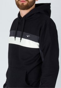 Hombre con barba que lleva una sudadera negra con rayas horizontales grises y blancas y un bolsillo frontal, de pie contra un fondo claro.
