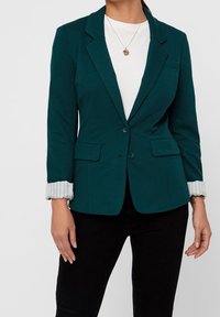 Teal colbert met lange mouwen, voorzien van twee voorkruiszakken en een knoopsluiting, gelaagd over een witte top met een gestreepte manchetdetail.