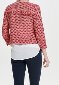 Rode en witte geblokte jas met franjes aan de achterkant, lange mouwen en een vierkante vorm, gelaagd over een wit shirt en donkere broek.