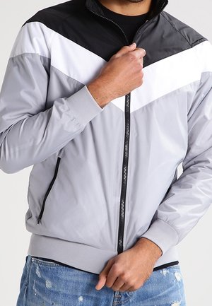 Homme portant une veste grise avec un motif chevron noir et blanc, en train de la fermer, assortie à un jean bleu déchiré.