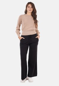 Donna con capelli lunghi, che indossa un maglione beige, pantaloni neri a gamba larga e sneakers bianche, in piedi con le mani in tasca su uno sfondo neutro.