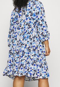 Bloemenjurk met een lichte achtergrond en blauwe, marineblauwe en zwarte bloemen. Gerimpelde zoom en 3/4 mouwen. Zacht stof met een vloeiende design.