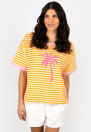 Woman with long dark hair wearing an orange and white striped T-shirt with a pink palm tree design, paired with white shorts, smiling slightly.