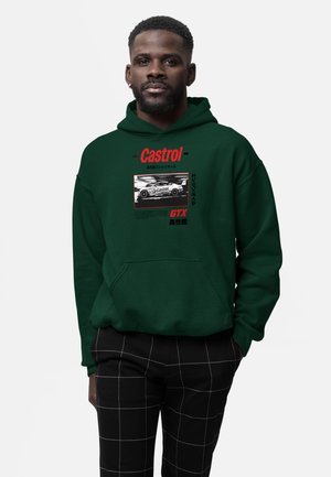 Homme portant un sweat à capuche vert foncé avec une voiture de course blanche et le texte rouge « Castrol GTX », associé à un pantalon noir à carreaux.