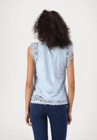 Ljusblå spetsblus med korta, scallopade ärmar, intrikata blommönster och en texturerad finish, i kombination med mörka denimjeans.