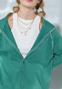 Sudadera verde con cremallera y una camiseta sin mangas blanca de canalé debajo. La sudadera presenta una cremallera plateada, bolsillos laterales y ribetes en contraste. Collares de oro en múltiples capas.
