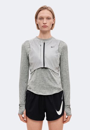 Une femme pose en tenue de sport grise, avec un gilet argenté zippé sur le devant et un short noir Nike, sur un fond uni.