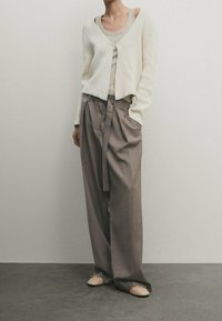 Weißer, strukturierter Cardigan über einem beigen Tanktop; lockere, graue Hose mit Kordelzug; hellfarbene flache Schuhe. Schlichtes, modernes Design.