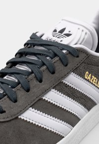 Grå mockasinsneaker med tre vita ränder, guldfärgad text "GAZELLE", grå snören och en vit tunga med Adidas-logotypen.