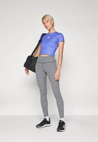 Ung person i blå crop top, grå leggings, svarte joggesko, som bærer en svart skuldertreningstask, stående mot en enkel, lys bakgrunn.