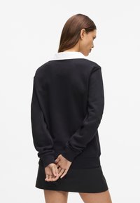 Sudadera negra con cuello blanco, que presenta un corte relajado y mangas largas. Textura suave y puños acanalados. El modelo se encuentra de pie con las manos detrás de la espalda.