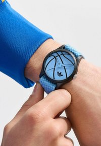 Melna pulksteņa ar zilu basketbola motīvu ciparnīcā, kurai ir teksturēts zils austs siksniņa. Pulksteņa rādītāji ir gaiši zili, ar Adidas logotipu.