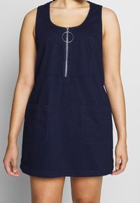 Nainen, joka on pukeutunut ilman hihoja olevaan tummansiniseen farkkuasuunsa, jossa on edessä vetoketju ja kaksi etutaskua, seisoo vaaleanharmaata taustaa vasten.