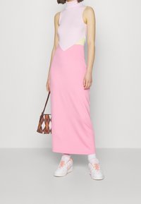 Różowa maxi sukienka o dopasowanym, bezrękawnikowym fasonie oraz stójce. Warstwowy, pastelowy top z geometrycznym wycięciem. Dodatki to wzorzysty torebka i sneakersy.