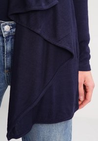 Mörkblå topp med ett lager-på-lager, draperad design, gjord av mjukt tyg. Har en texturerad yta och långa ärmar, matchad med denimjeans.