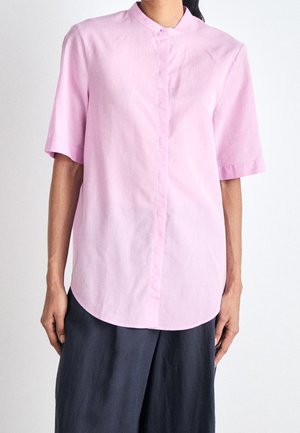 Persoon met een lichtroze korte mouwen blouse met knopen en een mandarijnkraag en donkere, wijde broek tegen een effen achtergrond.