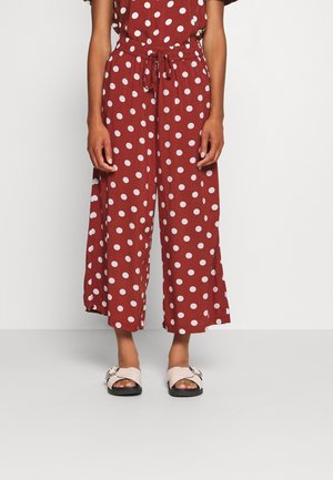 Femme portant un pantalon large couleur rouille avec des pois blancs, associé à des sandales beiges à enfiler avec de grandes boucles.