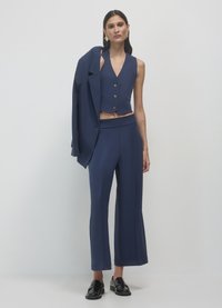 Gilet sartoriale blu navy con tre bottoni, pantaloni a vita alta abbinate con gamba svasata e blazer appoggiato sulla spalla. Mocassini neri.