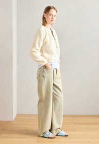 Casaco de pele texturada em creme claro, abotoado, combinado com uma camisa azul clara e calças largas em bege. As sandálias são azuis claras com correias ajustáveis.