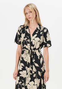 Robe noire avec imprimé floral surdimensionné en crème. Manches courtes, col et taille ceinturée avec une jupe fluide.