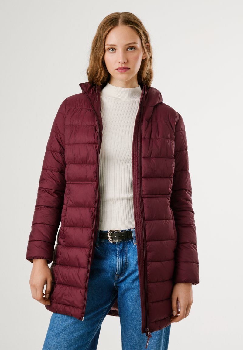 Bordo puffer jakna s patentnim zatvaračem sprijeda i kapuljačom. Ima pufast, kestenjast oblik i duge rukave. Noseća preko krem pulovera s rebrastim uzorkom.