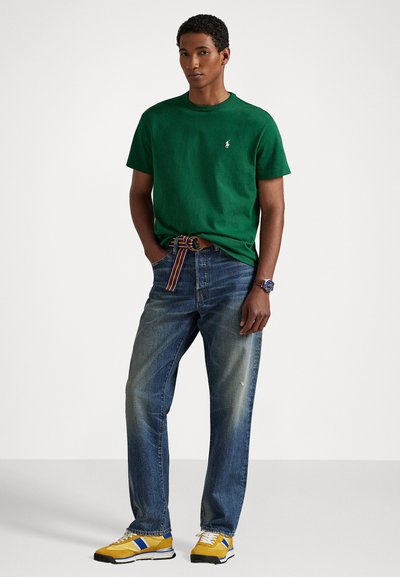 Camiseta de algodón verde, de cuello redondo, combinada con jeans azul desgastados. Un cinturón a rayas colorido y zapatillas amarillas con acentos azules completan el look.