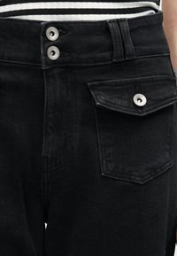 Zbliżenie na czarne dżinsy z podwójnymi srebrnymi guzikami i kieszenią z klapką po prawej stronie, noszone z czarno-białym paskowanym topem.