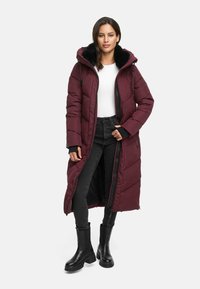 Cappotto lungo bordeaux imbottito con cappuccio in pelliccia sintetica, chiusura con zip, tasche laterali e motivo a quadri. Indossato sopra una camicia bianca e jeans neri.