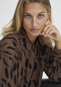 Brun bluse med sorte abstrakte mønstre, blødt stof, krave og knaplukning ved håndleddene. Modellen har langt hår og hviler hånden på hagen.