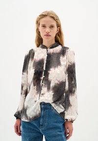 Bluza z dolgimi rokavi iz svile, ki ima črno-belo tie-dye vzorčenje z navpičnimi črtami, mandarin ovratnik in gumbasto zapiranje.