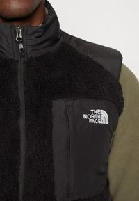 Homme portant une veste polaire noire avec zip et logo The North Face sur la poitrine, par-dessus une chemise à manches longues vert olive.