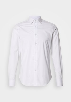 White long-sleeve shirt with small navy polka dots, button-down collar, and subtle sheen, featuring a curved hem and standard button front.