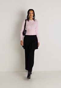 Ανοιχτό ροζ ριμπ μπολέρ με κουμπιά, συνδυασμένο με μαύρη πλισέ φούστα και μαύρα παντελόνια. Φορεμένο με μαύρες μπότες μέχρι τον αστράγαλο.