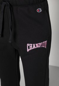 Černé tepláky s elastickým pasem, šňůrkou a vyšívaným logem "CHAMPION" v růžové barvě na levém stehně. Měkká texturovaná látka.