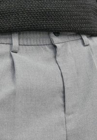 Šedé kalhoty na míru z hladkého materiálu, s hlubokými záhyby, zapínáním na knoflík a plochým pasem, který je vidět nad strukturovaným tmavým svetrem.