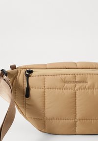 Quilted tan shoulder bag with a textured finish, black zipper, and adjustable strap. Features branded logo embossed on the surface.