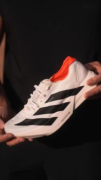 Chaussure de sport blanche avec des rayures noires, un accent orange sur le col et une semelle texturée. Présente un design à lacets et une languette rembourrée.