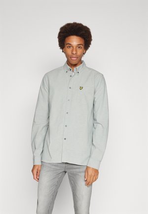 Camicia grigia chiara a maniche lunghe con colletto, con un piccolo logo giallo ricamato sulla tasca del petto, abbinata a jeans grigi chiari.