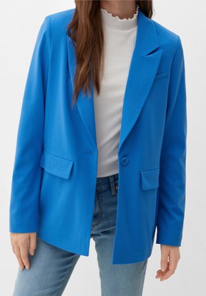 Frau trägt einen leuchtend blauen Blazer über einem weißen gerippten Oberteil und hellblauen Jeans, steht vor schlichtem Hintergrund.