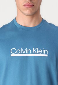 Modrá bavlnená tričko s bielym logom "Calvin Klein", okrúhlym výstrihom a krátkymi rukávmi, s hladkou textúrou a neformálnym dizajnom.