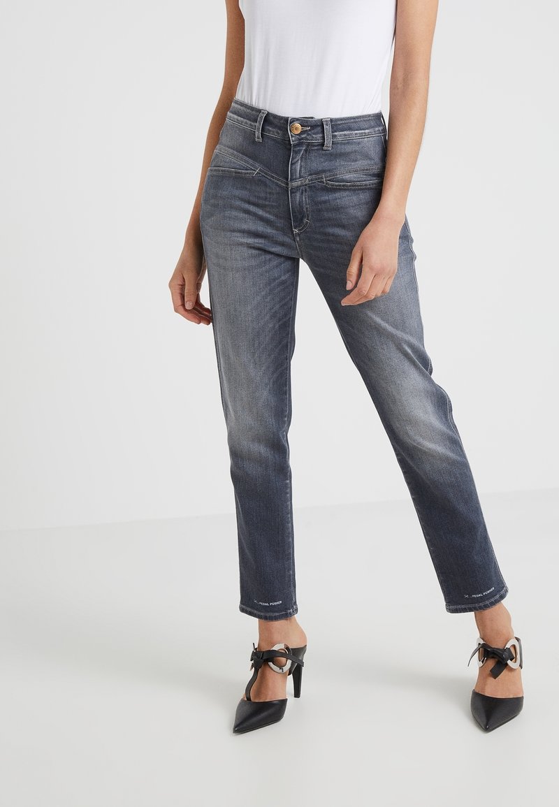 Femme portant un jean droit bleu foncé délavé associé à des chaussures à talons noirs ornées de détails en nœud et anneau.