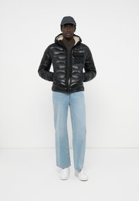 Zwarte pufferjack met ritssluiting, capuchon en zakdetails, gecombineerd met lichtblauwe spijkerbroek en witte sneakers. Eenvoudige, gladde textuur.