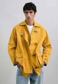 Chaqueta de algodón amarilla con collar puntiagudo, presenta cierres de tipo toggles, bolsillos laterales y puños en contraste, llevada sobre una camisa blanca.