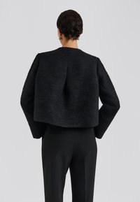 Krátká černá texturovaná bunda s dlouhými rukávy, vysokým límcem a detailními švy, doplněná o černé upravené kalhoty.